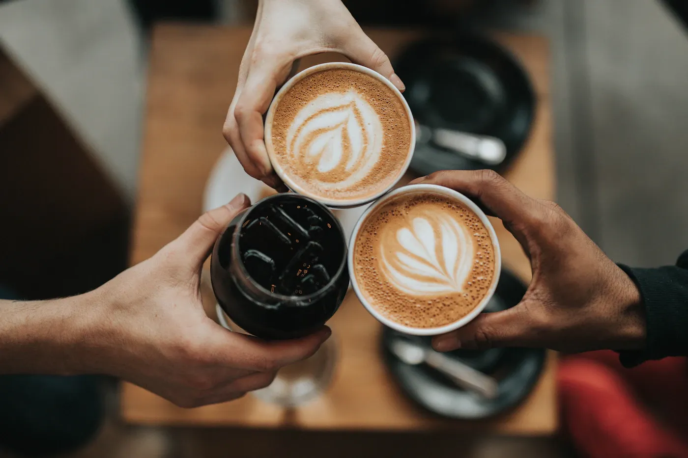 Drei Hände stoßen mit zwei Milchkaffees und einem kalten Kaffee an